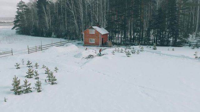 Aerial view. A Caucasian man holding an axe, chopping wood in winter. A man stands in the courtyard of a country house, which is located next to the forest. The camera flies towards the house.