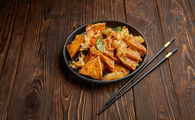 Homemade tofu stir fry with vegetables in hot sweet sauce and pair of chopsticks on dark wooden background and copy space. Asian food concept top view