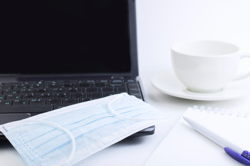 Medical mask on a laptop next to a coffee mug, and a ballpoint pen for notes. The concept of working at home business, home schooling, shopping. Work during a viral outbreak. Insulation.