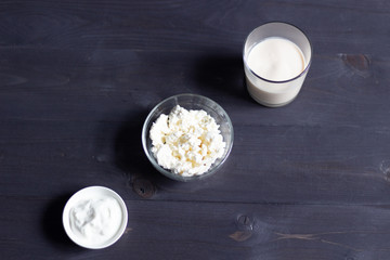 Homemade cottage cheese in bowl, glass of fermented baked milk, sour cream on black wooden background