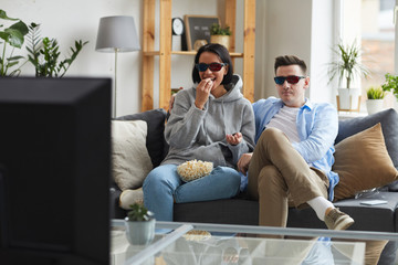Obraz premium Happy young couple wearing 3D glasses sitting on sofa and eating popcorn while watching the movie on TV