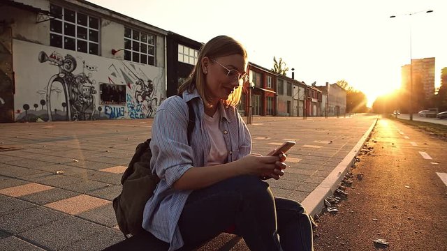 Smiling girl sitting on long board in the city during sunset looking at phone.
