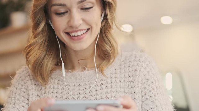 A laughing nice blonde young woman in wired earphones is looking to her smartphone holding it horizontally while drinking coffee in the white cozy cafe