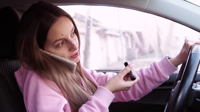 A young girl in pink clothes speaks on the phone and paints her lips while sitting in a car.