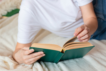 A young woman in a white T-shirt is reading a book lying on the bed. Home leisure - reading a book.
