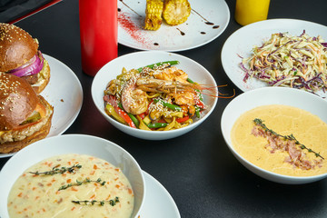 Assorted different dishes on a black background. Dining table with burgers, cream soups, wok noodles, cole slaw salad and grilled corn. Top view, food flat lay