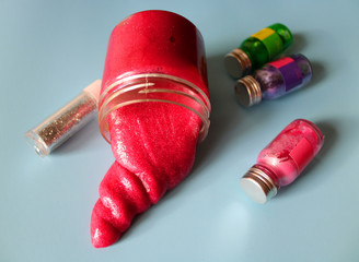 Pink slime flows out of a containernext to small jars of colored paints and glitters on a blue...