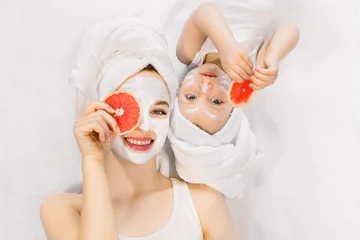 Fotobehang Pretty young mother and her little two years old daughter with towels on heads, lying together on the bed and doing spa care skin procedures, facial mask, holding pieces of grapefruit near their eyes © sofiko14