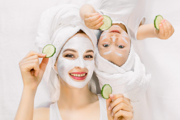Mother and daughter in white shirts and white towels on their heads in a home bathroom, doing spa procedures with mud mask and fresh cucumber slices. Family time, spa and beauty, mothers day
