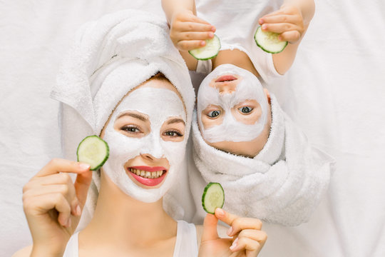 Happy Loving Family, Mother And Little Baby Girl, With Towels On Their Heads, Are Doing Mud Facial Mask, Holding Cucumer Slices And Having Fun Lying On Isolated White Background. Family Spa