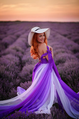 Happy young red-haired woman in luxurious dress standing in lavender field at sunset