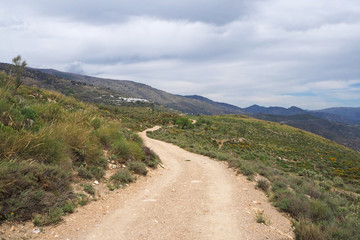 The landscape with the slope covered by green grass, big yellow ground way, far village with white houses, cloudy sky