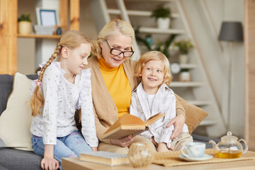 Mature woman in eyeglasses sitting on sofa together with her grandchildren and reading a book to them at home