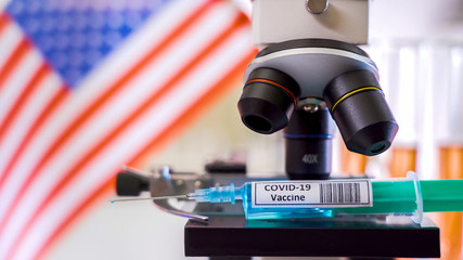 The USA flag waving on the back of the microscope with the coronavirus syringe