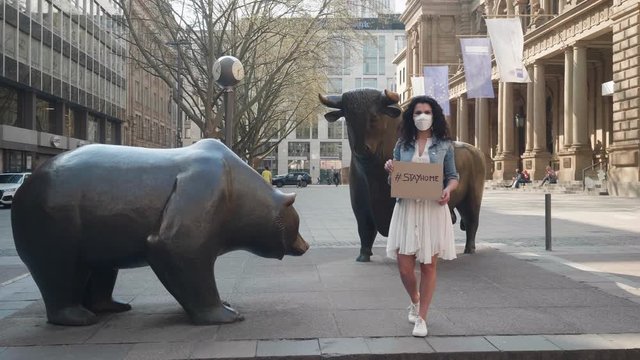 Frankfurt, Germany, April 8, 2020 Young women waering protective face mask against corona covid-19 virus holding sign with #stayhome in front of bull and bear stock market exchange shot in 4k