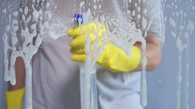 Woman wearing rubber glove spraying soap on window glass cleaning it with squeegee