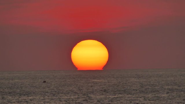 huge sun, sunset into sea water with orange colorful twilight sky background, silhouette small couple sitting on board watching sunset in middle ocean