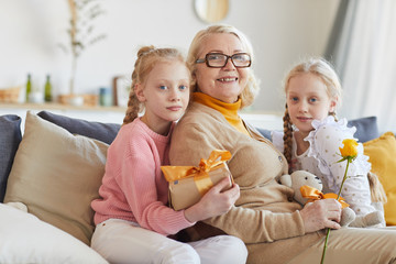 Portrait of happy grandmother in eyeglasses sitting on sofa together with her granddaughters and they smiling at camera