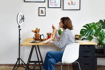 Female artist filming design workshop at home for online education. Working from home making master-class using ring lighting. Cozy artist's studio.