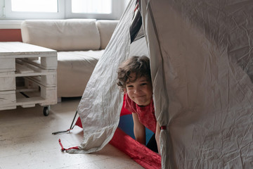 Indoor life at the coronavirus quarantine. Cute curly boy looks from a tent at home.