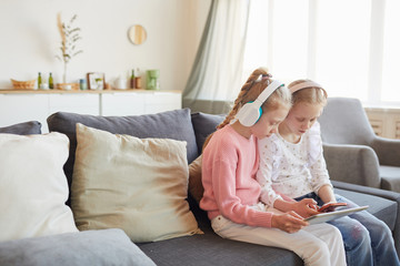 Two sisters in headphones sitting on sofa with digital tablet and playing together in online games...