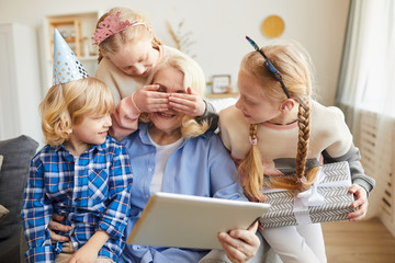 Grandmother sitting in armchair and using tablet pc while her grandchildren making a surprise for...