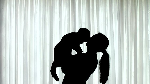Silhouette of woman lifting a baby up in front of a window with white curtains