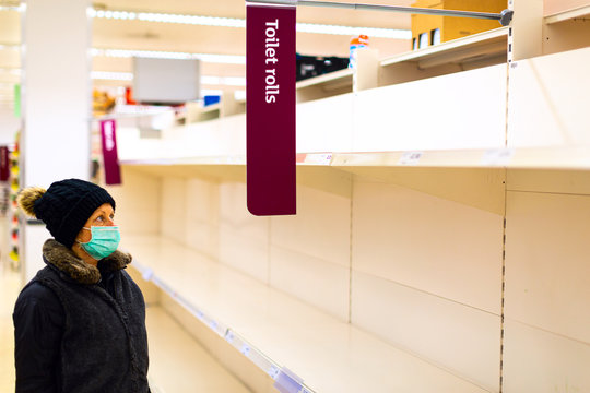 Senior Female Customer Wearing A Protective Medical Mask Looks Confused At The Empty Shelves Of Toilet Paper Rolls In A Supermarket