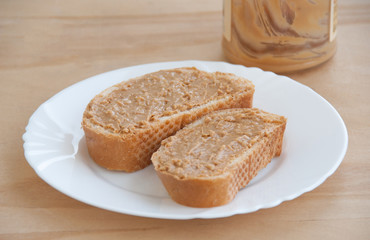 Peanut butter toast on a white plate in front of a jar. Wooden background. Close up