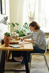 Fototapeta na wymiar Female artist at her workplace working from home. Woman dressed in jeans and striped shirt sitting at the table and drawing illustration.