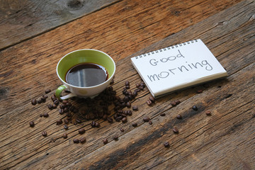 Fresh Cup of brewed coffee on a wooden background and a greeting note in the notebook Good Morning 