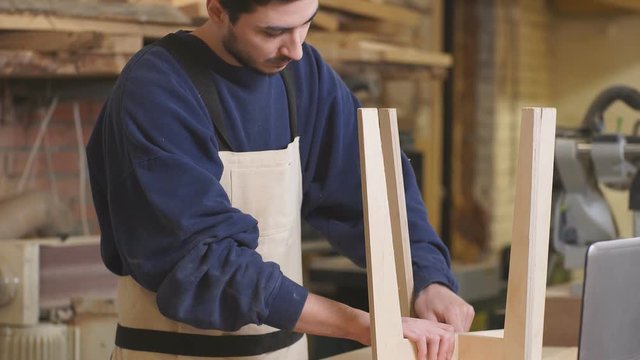 Serious caucasian carpenter deals with making wooden chair in workshop, construction, industry concept.