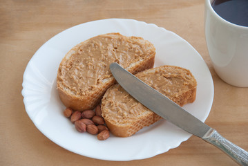 peanut butter sandwich on a white plate. White cup of coffee, knife and peanuts. Close up