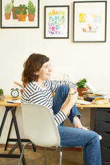 Female artist at her workplace working from home. Young woman dressed in jeans and striped shirt sitting at the table, smiling and creating an illustration.