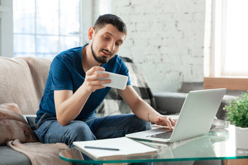 Young focused man studying at home during online courses or free information by hisself. Becomes economist, financist, translator while isolated, quarantined. Using laptop, smartphone, headphones.