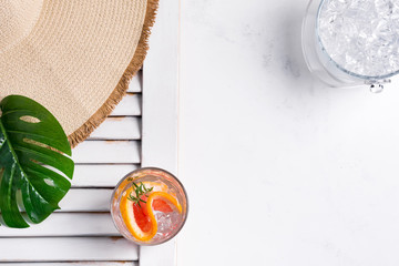 Cold summer drink in a glass with slice of grapefruit and ice cubes and Wide-brimmed hat and green tropical plant leaf on a white background with part of wooden jalousie. Top view.