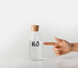 A hand points a finger at a glass bottle with the chemical formula of water on it and a clear white liquid inside.