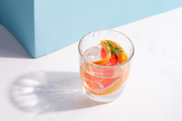 Refreshing cold summer drink in a glass with slice of grapefruit and ice cubes on a white table with shadow against pastel blue wall.
