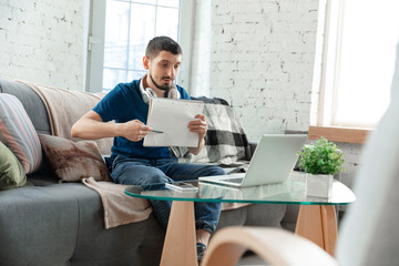 Young focused man studying at home during online courses or free information by hisself. Becomes analyst, retailer, ingeneer while isolated, quarantined. Using laptop, smartphone, headphones.