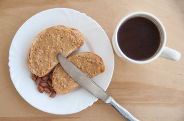 Coffee and peanut butter toasts on a white plate with a knife and peanuts. Wooden table. Top view