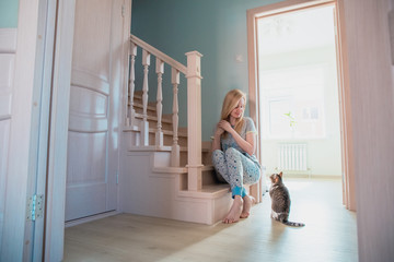 blonde girl sits on the bottom step of the stairs in a house with a bright interior and looks at the cat