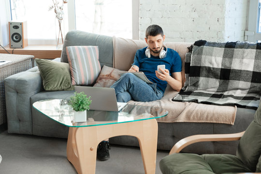 Young Focused Man Studying At Home During Online Courses Or Free Information By Hisself. Becomes Nurse, Constraction Laborer While Isolated, Quarantined. Using Laptop, Smartphone, Headphones.