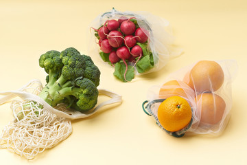 Zero waste food shopping with reusable bags. Flat lay with fruits and vegetables in textile packaging. Broccoli, radishes and oranges on yellow background.