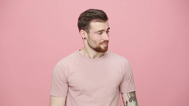 Confident strong handsome bearded tattooed young guy 20s in t-shirt isolated on pastel pink background studio. People emotion lifestyle concept Looking aside and at camera with charming smile laughing