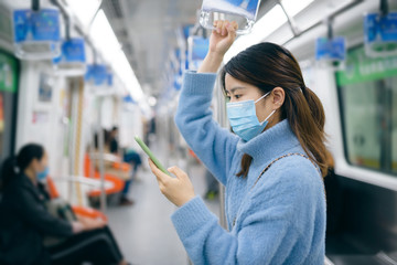 That young Asian woman wearing a surgical mask is using smart phone in the subway train.