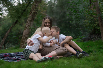 Naklejka na ściany i meble Young mom with three little children having picnic in summer. Big cousin hug and kiss her younger sister.