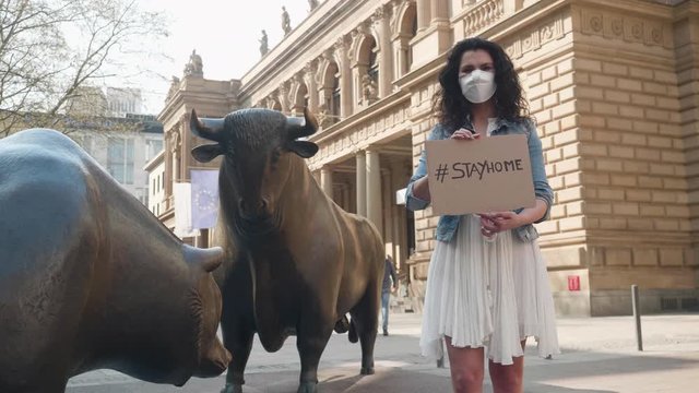 Frankfurt, Germany, April 8, 2020 Young women waering protective face mask against corona virus holding sign with COVID-19 and #stayhome in front of bull and bear stock market exchange shot in 4k