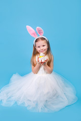 Obraz na płótnie Canvas Cute positive little girl wearing bunny ears holding easter rabbit on blue background.
