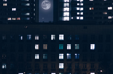 Landscape of a night city with high-rise buildings and light in the windows of residents and a full moon between the houses