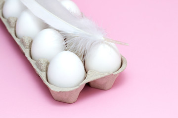 White Easter eggs in a cardboard box with chicken feather on a pink background